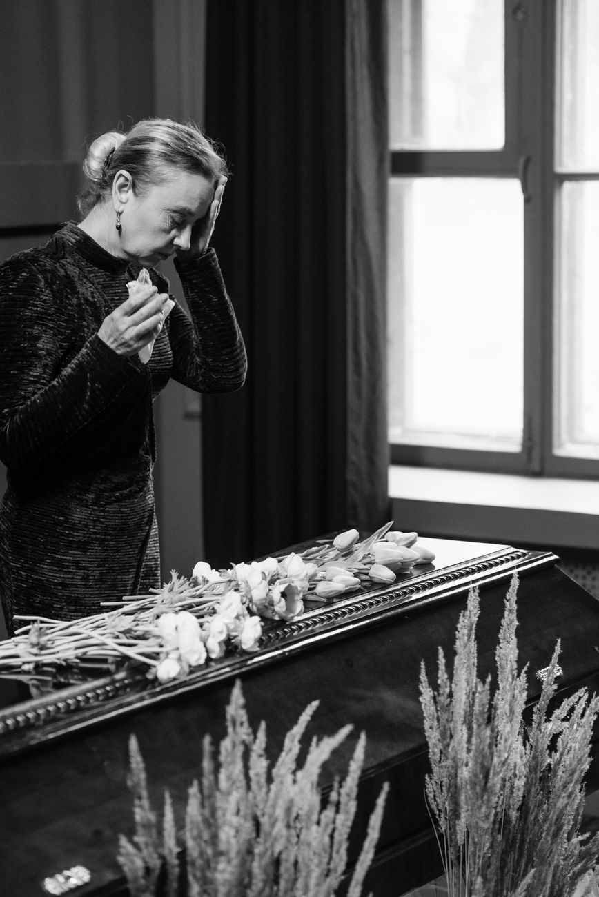 Home Woman mourning at a funeral, standing beside a coffin, holding flowers indoors.