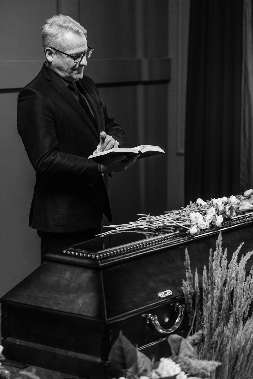 Home Black and white image of a pastor reading the Bible at a funeral service with a coffin and flowers.