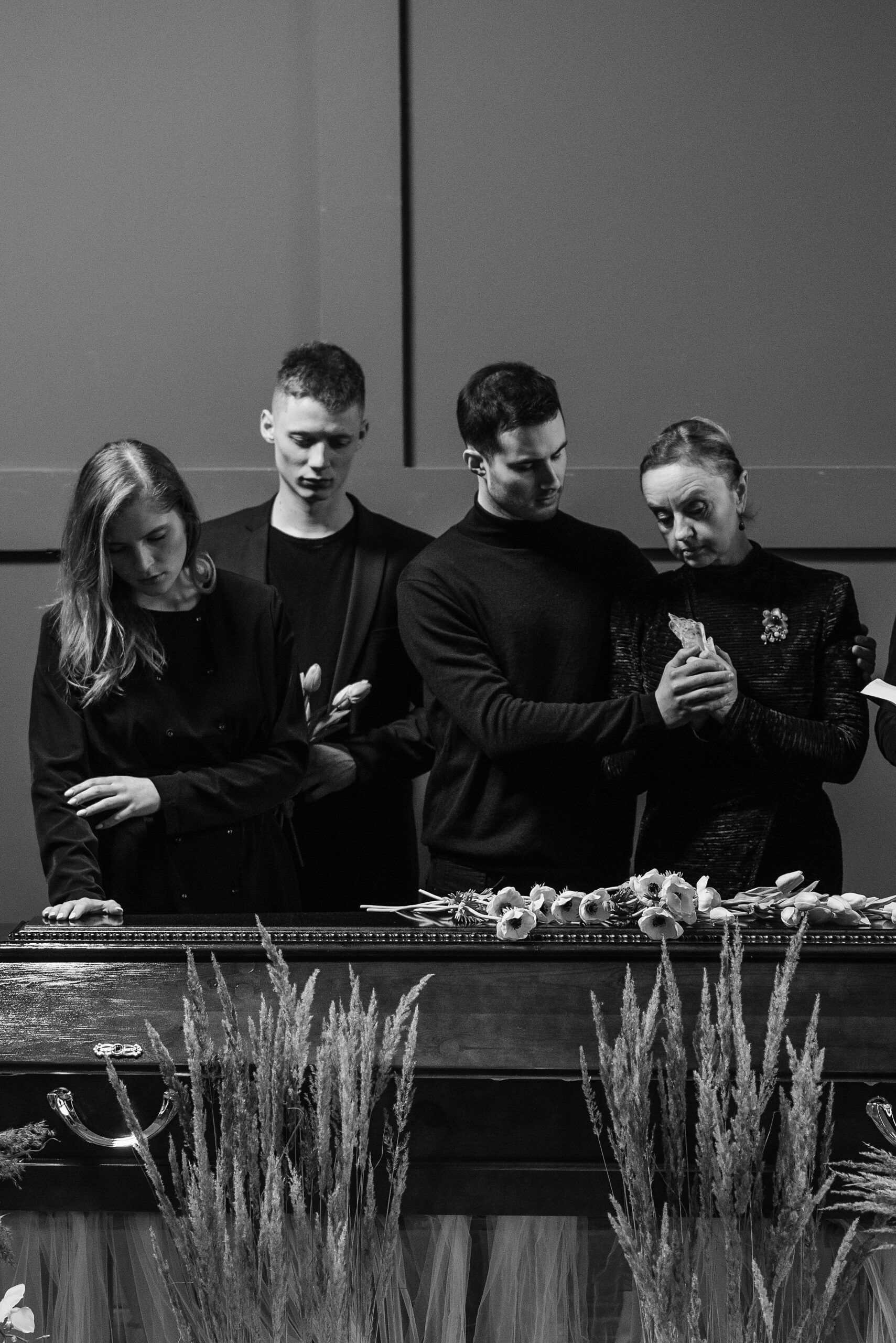 Home A grayscale image of a family mourning at a funeral service, standing by a coffin with flowers, expressing sorrow and loss.